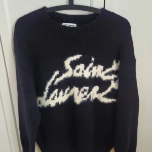 Svart tröja från Saint Laurent - Snygg svart tröja från Saint Laurent med vit text på framsidan. Tröjan har långa ärmar och en ribbad rund halsringning. Perfekt för en stilren look. Oanvänd, lappen sitter kvar. Inget kvitto färsv bra pris.