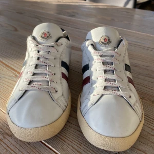 Vita sneakers från Moncler - Snygga vita sneakers från Moncler med klassiska ränder i rött och blått på sidan. Skorna har snörning och en krämfärgad sula som ger en retro känsla. Perfekta för en stilren look.                                        Skicket är väldigt bra men man bedömer skick själv😀skulle själv säga 7,5/10 i skick                                       Självklart äkta men tyvärr inget OG med               Hör av er vid minsta lilla!     Billigare vid snabb affär! Skicka gärna byten också!😀(liten repa i bak!!)