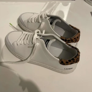 Vita sneakers med leoparddetaljer - Snygga vita sneakers från Axel Arigato med leopardmönstrade detaljer på hälen. Skorna har snörning och en rund tå. Perfekta för att ge din outfit en trendig touch.
