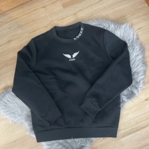 Svart sweatshirt från Shein med vingar - Säljer en svart sweatshirt från Shein med tryck av vita vingar och texten 'ANGEL' på bröstet. Tröjan har rund halsringning med textdetalj och långa ärmar. Perfekt för en avslappnad stil.