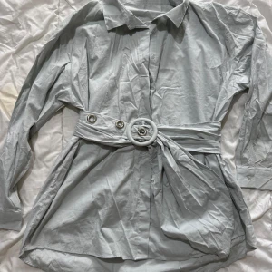 Ljusgrå skjorta med bälte - Snygg ljusgrå skjorta med knappar och ett matchande bälte i midjan. Skjortan har långa ärmar och en klassisk krage. Perfekt för en stilren look.