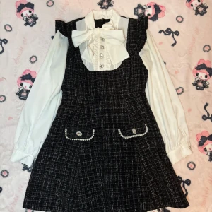 Svart och vit klänning med rosett från Ank Rouge - Elegant svart och vit klänning från Ank Rouge med en stor vit rosett och dekorativa knappar framtill. Klänningen har långa vita ärmar och fickdetaljer med pärlor. I tweed, dragkedja på ryggen. Nån fläck på ena armen 