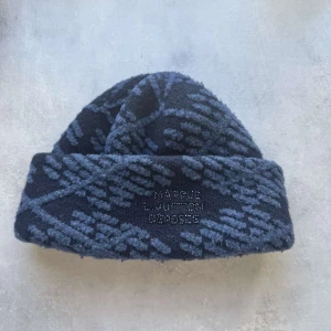 Louis Vuitton mössa - Snygg och trendig mössa från Louis Vuitton med ett unikt damier argyle-mönster. Den är i en mörkblå nyans med ljusare blå detaljer och har en broderad logotyp framtill. Perfekt för att hålla sig varm och stilren under kyliga dagar.