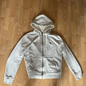 Grå hoodie från Polo Ralph Lauren - Säljer en stilren grå hoodie från Polo Ralph Lauren. Den har en dragkedja framtill och en klassisk logga på bröstet. Perfekt för en avslappnad look. Kan gå ner i pris vid en snabb affär.