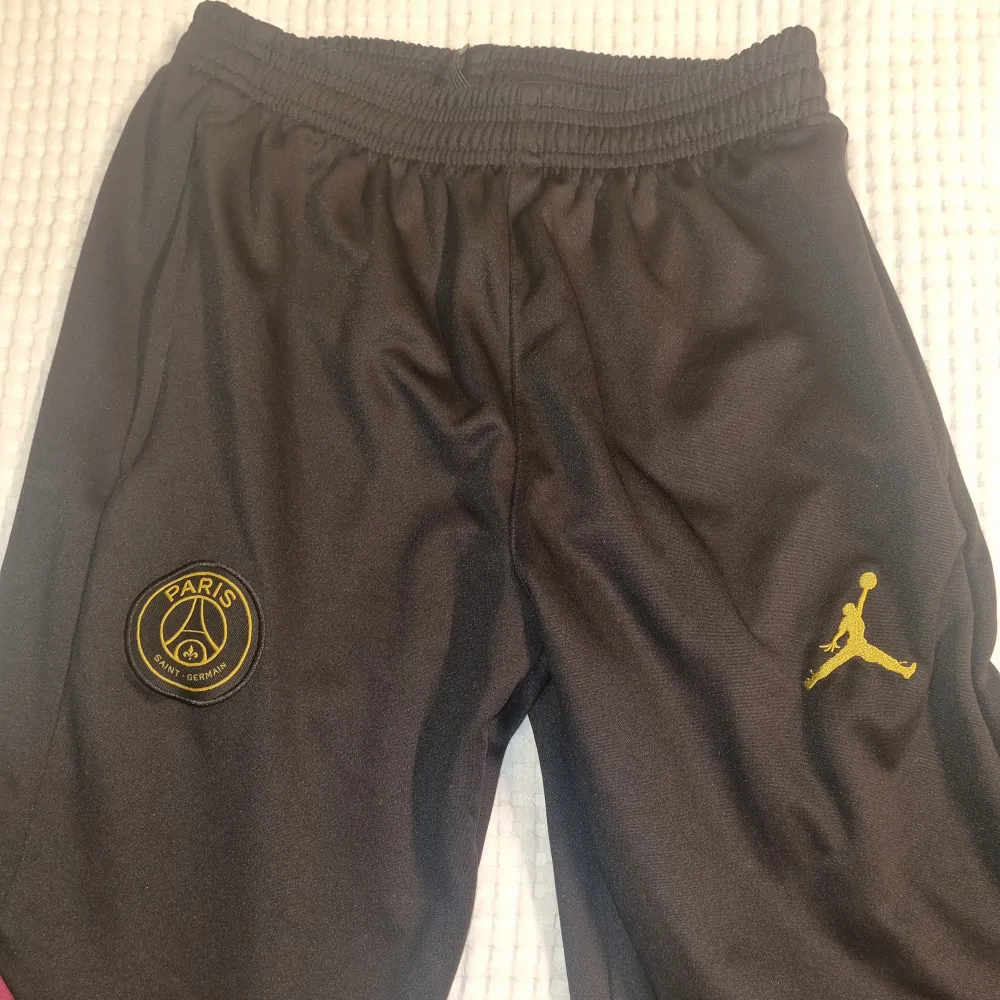 Svarta träningsbyxor med PSG-logga och guldfärgad Jumpman. Byxorna har elastisk midja och rosa detaljer längs benen. . Farkut & Housut.