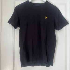 Svart t-shirt från Lyle & Scott - Säljer en klassisk svart t-shirt från Lyle & Scott med deras ikoniska gula logga på bröstet. Perfekt för en stilren och enkel look. T-shirten är kortärmad och gjord i mjukt material.