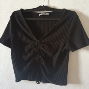 Svart ribbad topp från FB Sister - Säljer en svart ribbad topp från FB Sister med korta ärmar och en snygg knytdetalj framtill. Toppen har en v-ringad design och är perfekt för en stilren look.