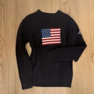 Svart/mörkblå tröja med amerikansk flagga från Gant - Säljer en svart/mörkblå tröja från Gant med en stor amerikansk flagga på framsidan. Tröjan har långa ärmar och texten 'GANT NEW HAVEN 1949' på ärmen. Perfekt för en stilren och casual look.