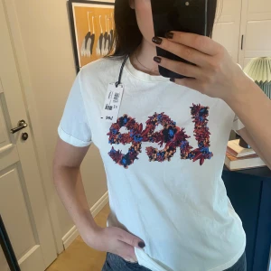 Vit t-shirt från Lee med blommigt tryck - Säljer en vit t-shirt från Lee med ett blommigt tryck i rött och blått som formar märkesnamnet. Perfekt för en avslappnad stil. T-shirten är kortärmad och gjord i 100% bomull.