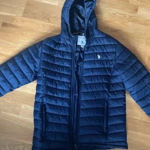 Mörkblå jacka från U.S. Polo Assn. - Säljer en mörkblå jacka från U.S. Polo Assn. med dragkedja och huva. Jackan har ett quiltat mönster och är perfekt för våren . Den har en broderad logga på bröstet och är långärmad. Den är använd ett fåtal gånger men i bra skick.