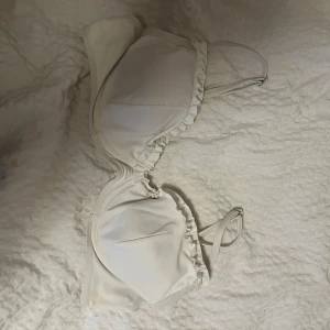 Vit bikiniöverdel med volangdetaljer - Säljer en söt vit bikiniöverdel med volangdetaljer längs kanterna. Den har smala axelband och en mjuk, bekväm passform. 