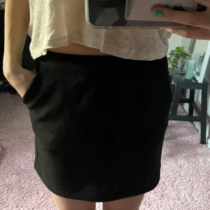 Svart kortkjol med dragkedja - Snygg svart kortkjol i mocka med dragkedja bak. Perfekt för en stilren look. Kjolen har en enkel design som gör den lätt att matcha med olika toppar.