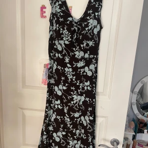 Brun/svart blommig klänning - Snygg brun klänning med ljusblått blommönster. Den har en v-ringad design och knytning framtill. Perfekt för en somrig look!