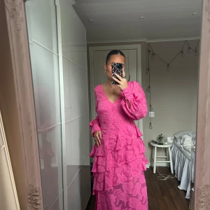 Rosa långklänning med volanger - Rosa långklänning med volanger och blommigt mönster. Klänningen har en v-ringad design och långa ärmar. Materialet är lätt och luftigt, vilket gör den bekväm att bära. Den har ett väldigt lite hål precis vid dragkedjan som inte alls syns när klänningen är på. Använd 1 gång. Köpt på Asos för 1400kr.