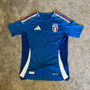 Blå fotbollströja från Adidas - Säljer en blå fotbollströja från Adidas med Italiens landslagsemblem på bröstet. Tröjan har korta ärmar och ett stilrent mönster med vita och gröna detaljer på axlarna. Perfekt för fotbollsfans! 💙