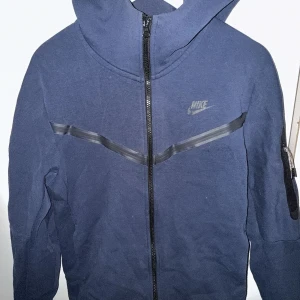 Mörkblå Nike tech fleece i från Nike - Säljer en mörkblå jacka från Nike med dragkedja och huva. Jackan har en stilren design med diskret Nike-logga på bröstet och en praktisk ficka på ärmen. Perfekt för en sportig look.