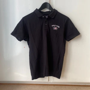 Svart pikétröja från Polo Club - Snygg svart pikétröja från Polo Club med klassisk krage och knappar framtill. Perfekt för en stilren look. Diskret logga på bröstet.