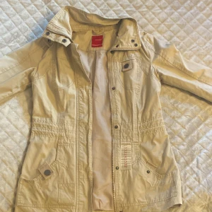 Beige jacka från Vero Moda - Snygg beige jacka från Vero Moda med dragkedja och knappar. Jackan har en normal passform och är perfekt för vår och sommar. Den har långa ärmar och praktiska fickor framtill.