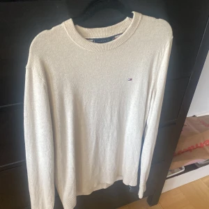 Beige stickad tröja från Tommy Hilfiger - Säljer en stilren beige stickad tröja från Tommy Hilfiger med rund halsringning och diskret logga på bröstet. Perfekt för lager-på-lager och passar till det mesta. Långa ärmar och ribbade muddar.