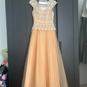 Champagnefärgad långklänning med spetsdetaljer - Säljer en elegant långklänning i champagnefärg med vackert blommigt spetsliv och lätt utställd kjol i tunt material. Klänningen har v-ringning och små ärmar, perfekt för bal eller fest. Blommönster och transparenta detaljer upptill ger en lyxig känsla.