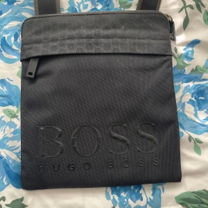 Svart axelväska från Hugo Boss - Snygg svart axelväska från Hugo Boss med stort präglat logotyptryck på framsidan. Väskan har dragkedja upptill och justerbar axelrem. Perfekt för att bära med sig det viktigaste. Köpt ifrån Hugo Boss för 1200kr 
