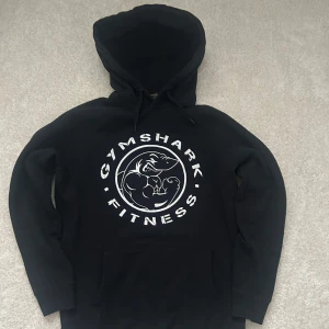 Svart hoodie från Gymshark - Säljer en svart hoodie från Gymshark med stort vitt tryck på bröstet där det står 'GYMSHARK FITNESS' och en muskulös haj i mitten. Tröjan har huva och magficka, perfekt för gymmet eller chill.