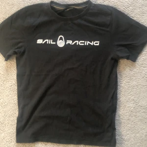 Svart t-shirt från Sail Racing - Svart t-shirt från Sail Racing med vit logotyp och text på bröstet. Klassisk rund halsringning och korta ärmar. Perfekt för en sportig och stilren look.