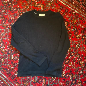 Svart långärmad tröja från Selected Homme - Säljer en svart stickad långärmad tröja från Selected Homme. Tröjan är tillverkad i 100% ekologisk bomull och har en enkel, stilren design med rund hals. Perfekt för en clean och avslappnad look.