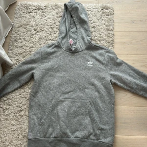 Grå hoodie från Adidas - Säljer en klassisk grå hoodie från Adidas med vit broderad logga på bröstet. Tröjan har huva och en stor magficka framtill. Knappast använd och jätte bra skick 