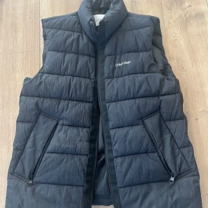 Svart dunväst från Calvin Klein - Snygg svart dunväst från Calvin Klein med diskret logga på bröstet. Västen har ståkrage, två fickor med dragkedja och quiltad design. Tillverkad med DuPont Sorona för lätt och varm känsla.