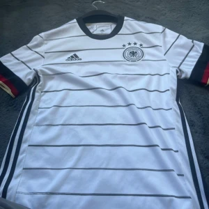 Tysklands landslagströja från Adidas - Vit fotbollströja från Adidas med svarta horisontella ränder och Tysklands landslagsemblem på bröstet. Tröjan har svarta detaljer vid krage och ärmslut samt röda och gula inslag på ärmarna. Tillverkad i lätt och ventilerande Aeroready-material.