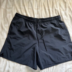Svarta träningsshorts från Nike - Svarta träningsshorts från Nike med elastisk midja och snörning. Shortsen har två sidofickor och är tillverkade i ett lätt och snabbtorkande material, perfekt för träning eller sport. Diskret Nike-logga broderad på ena benet. Passar xl och 2xl!