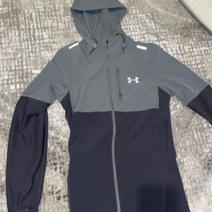 Grå och svart träningsjacka från Under Armour - Säljer en snygg grå och svart träningsjacka från Under Armour med huva och dragkedja. Jackan har lång ärm och är perfekt för utomhusträning. Logotypen syns på bröstet och jackan har sportig design.