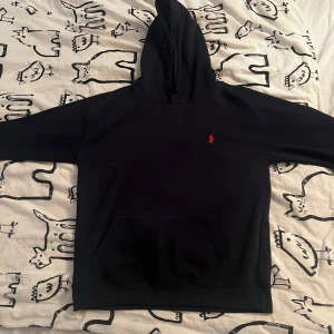 Svart hoodie från Polo Ralph Lauren - Säljer en mörkblå hoodie från Polo Ralph Lauren med klassisk röd broderad logga på bröstet. Tröjan har huva  och en stor magficka framtill. Har använt par gånger.Perfekt för dig som gillar stilrena och bekväma plagg.