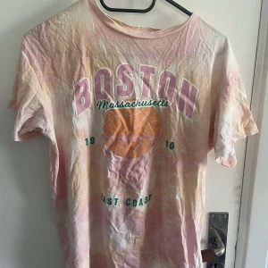 Pastellfärgad t-shirt med Boston-tryck från H&M - Säljer en pastellfärgad t-shirt från H&M med stort Boston-tryck och basketboll på bröstet. T-shirten har korta ärmar och en avslappnad passform. Perfekt för dig som gillar sportiga och färgglada plagg!