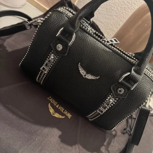 Zadig & Voltaire väska - Säljer en svart handväska från Zadig & Voltaire med silverfärgade detaljer och dekorativa dragkedjor. Väskan har ett emblem med vingar framtill och är tillverkad i strukturerat skinn. Perfekt accessoar för att lyfta din outfit. Den har en defekt annars i superbra skick. Skriv för bilder eller frågor.