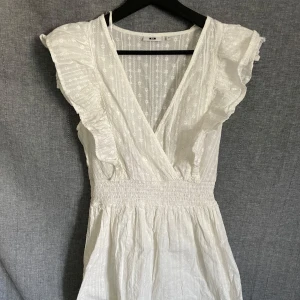 Vit omlottklänning med volangdetaljer - Superfin vit omlottklänning med v-ringning och volangdetaljer längs axlarna. Klänningen har resår i midjan och är ärmlös, perfekt för varma dagar. Tunt, lätt material med diskret mönster.