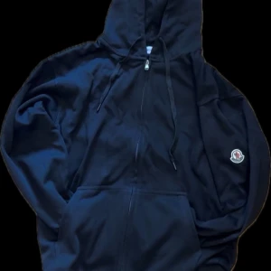 Svart hoodie från Moncler med dragkedja - Säljer en svart hoodie från Moncler med dragkedja framtill och huva med snörning. Jackan har en broderad Moncler-logga på ärmen och två fickor framtill. Perfekt för en avslappnad stil.