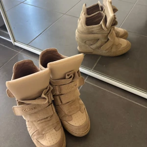 Isabel marant liknande skor - Säljer ett par beiga sneakers från Carinii med högt skaft och breda kardborreband. Skorna har en sportig design med rund tå och detaljer i mocka och syntet. Perfekta för dig som gillar streetstyle och vill sticka ut.