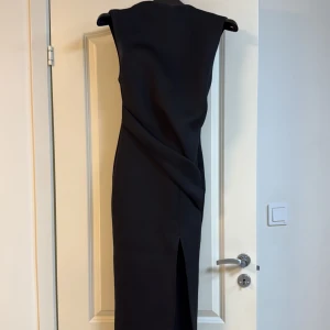 Svart långklänning med slits - Säljer en elegant svart långklänning med slits framtill och draperad detalj i midjan. Klänningen är ärmlös och har en snygg, figurnära passform. Perfekt för dig som vill ha en stilren och tidlös look. Tjockt tyg!! Väldigt bekväm 