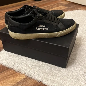 Svarta sneakers från Saint Laurent - Svarta sneakers från Saint Laurent med vit sula och broderad logga på sidan. Skorna har snörning och rund tå, samt en klassisk låg profil. Perfekta för dig som gillar stilrena och tidlösa sneakers. OBS (Hål vid hälen)