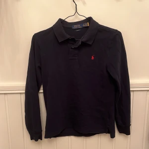 Mörkblå långärmad pikétröja från Polo Ralph Lauren - Säljer en mörkblå långärmad pikétröja från Polo Ralph Lauren med röd broderad logga på bröstet. Tröjan har klassisk krage och knäppning vid halsen. Slim fit-modell som passar perfekt till många tillfällen.