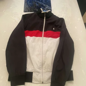 Ralph Lauren jacka  - Snygg vindjacka från Polo Ralph Lauren i svart, vitt och rött. Jackan har dragkedja framtill, ribbade muddar och klassisk logga på bröstet. Perfekt för en sportig och stilren look.