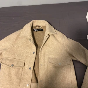 Beige  jacka från Zara - Säljer en beige overshirt jacka från Zara i storlek XS. Jackan har stora bröstfickor med knappar och klassisk krage. Perfekt för lager-på-lager och har en stilren look med tryckknappar framtill.