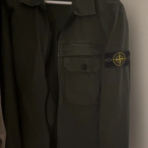 Grön overshirt från Stone Island - Säljer en grön overshirt från Stone Island med klassisk patch på vänster ärm. Skjortan har dragkedja framtill, bröstficka med knapp och en stilren krage. Perfekt för lager-på-lager och streetwear-stil. Storlek small men passar även medium skulle jag säga. 