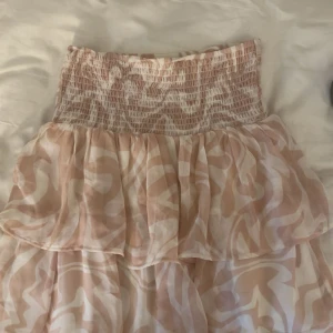 Beige och vit volangkjol med mönster - Säljer en söt kortkjol i beige och vitt med volanger och resår i midjan. Kjolen har ett abstrakt mönster och är gjord i ett lätt, luftigt material som passar perfekt till varma dagar. Använd fåtal gånger 