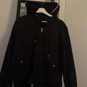 Svart vindjacka från Moncler - Säljer en svart vindjacka från Moncler med huva och dragkedja framtill. Jackan har snörning i midjan och två fickor med knappar. Klassisk Moncler-logga på ärmen och diskreta detaljer. Perfekt för blåsiga dagar. Tagg osv funkar Scan med 
