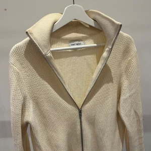 Beige kofta från EastWest - Säljer en beige stickad kofta från EAST WEST i storlek S. Tröjan har hel-zip, hög krage och är ribbstickad för en snygg och enkel look. Perfekt för lager-på-lager under svala sommarkvällar.