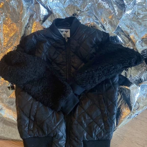 Svart quiltad bomberjacka med fuskpälsdetaljer - Säljer en svart quiltad bomberjacka med mjuka fuskpälsdetaljer på ärmarna. Jackan har dragkedja framtill och ribbade muddar vid ärmslut och nederkant. Perfekt för dig som vill sticka ut lite extra.