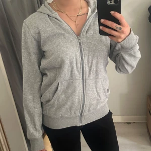 Grå hoodie med dragkedja - Säljer en klassisk grå hoodie med dragkedja och huva. Tröjan har långa ärmar, två fickor framtill och ribbade muddar vid ärmslut och nederkant. Perfekt för en avslappnad stil.